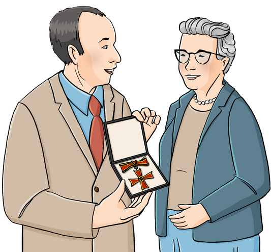 Ein Mann überreicht einer älteren Frau einen kreuzförmigen Orden in einer Schachtel. Beide tragen förmliche Kleidung.