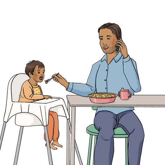 Ein Mann und ein Kleinkind sitzen am Esstisch. Das Kind sitzt im Hochstuhl und hat den Mund geöffnet. Der Mann hält ihm eine Gabel mit Nudeln vor den Mund. Gleichzeitig telefoniert der Mann mit dem Handy. Er schaut das Kind nicht an.