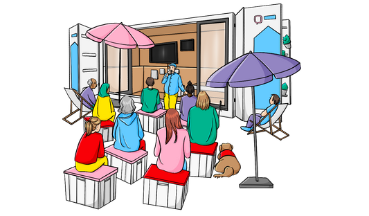 Menschen sitzen auf Kisten vor einem Container mit offenen Türen. Vor dem Container steht ein Mann und spricht in ein Mikro. Im Container hängt Bildschirm an der Wand.