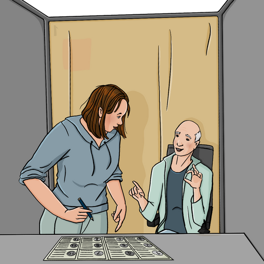 Ein Mann im Elektrorollstuhl in einer Wahlkabine. Neben ihm steht eine Frau mit einem Stift in der Hand. Sie schaut den Mann an. Der Mann zeigt auf den Stimmzettel, der auf dem Tisch liegt.