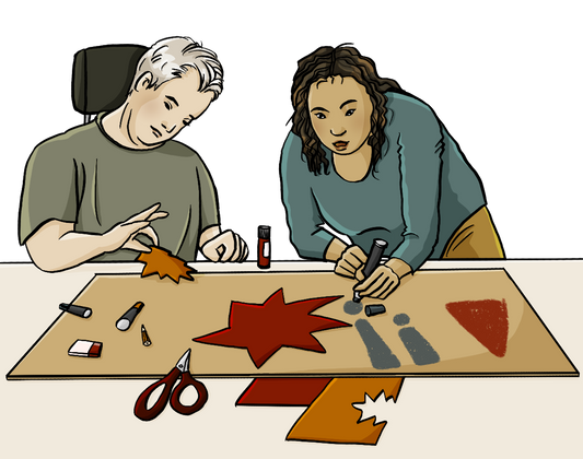 Ein Mann im Rollstuhl und eine Frau hinter einem Tisch. Auf dem Tisch liegen ein großes Stück Pappe und kleinere bunte Papierstücke, eine Schere, Stifte, ein Radiergummi und ein Klebestift. Die Frau schreibt gerade Ausrufezeichen auf das Stück Pappe, der Mann hält ein ausgeschnittes Stück Papier zwischen den Fingern.