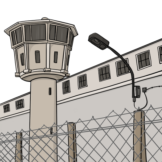 Ein Gebäude hinter einem Maschendraht-Zaun. Im Vordergrund steht ein Wachturm.