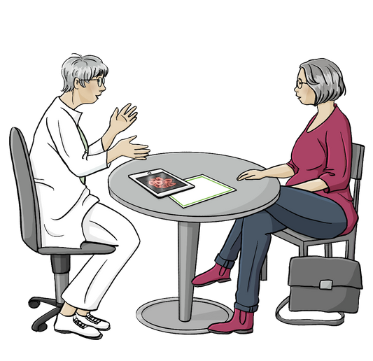 Zwei Frauen sitzen um einen Tisch. Eine von Ihnen trägt einen weißen Kittel und eine weiße Hose, die andere trägt Alltagskleidung. Auf dem Tisch liegt ein Tablett.