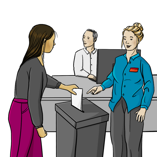 Eine Frau zeigt auf einen Wahlbehälter. Eine andere Frau steckt einen Zettel in den Behälter. Im Hintergrund sitzt ein Mann am Schreibtisch. 